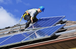 A worker in a blue hard hat installs solar panels on a roof under a blue sky.